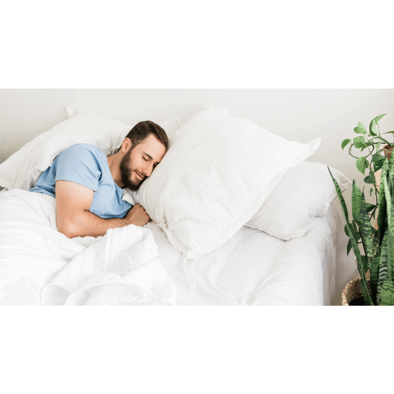 A imagem mostra um homem de camiseta azul, dormindo em uma cama com lençois brancos e travesseiros brancos. 