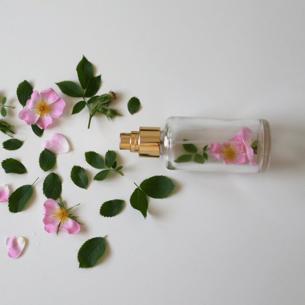 A imagem mostra um frasco de perfume transparente, dentro dele há algumas flores rosas e algumas folhas, elas também se espalham para fora do frasco.