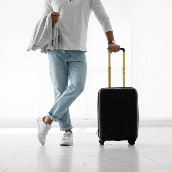 A foto mostra um homem com camiseta branca de manga comprida, calça jeans e tênis branco. Ele está apoiado em sua mala de viagem na cor preta.