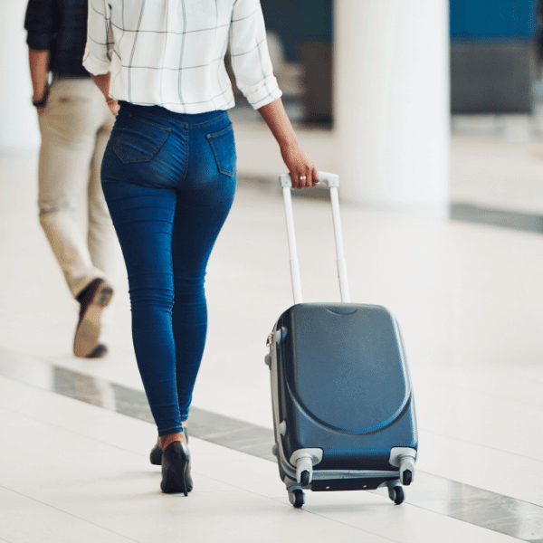 A foto mostra uma mulher de costas vestindo um blazer branco xadrez e uma calça jeans, ela está segurando uma mala de viagem.