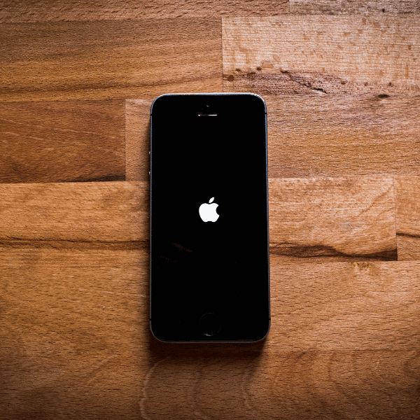 A foto mostra um iPhone em cima de um chão de madeira, sua tela está preta e mostra apenas o logotipo da Apple.