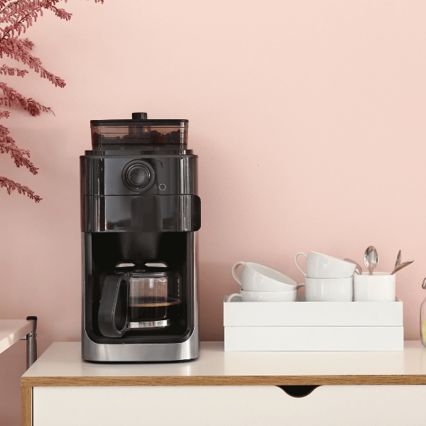 A foto mostra um cantinho do café composto por uma cafeteira elétrica e um kit de xícaras brancas.